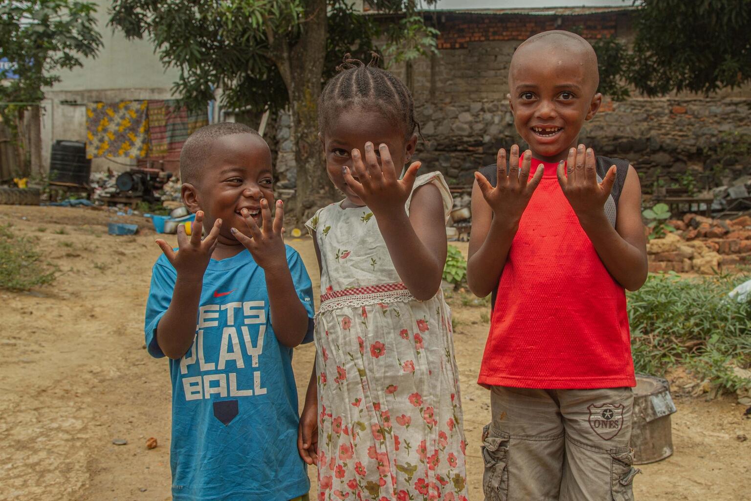 DRC - Children Displaying Ink Marks After Receiving Vaccinations Three young children standing outdoors proudly show ink marks on their fingers, indicating they've received vaccinations. They are smiling and appear playful, with a modest neighborhood setting in the background.
