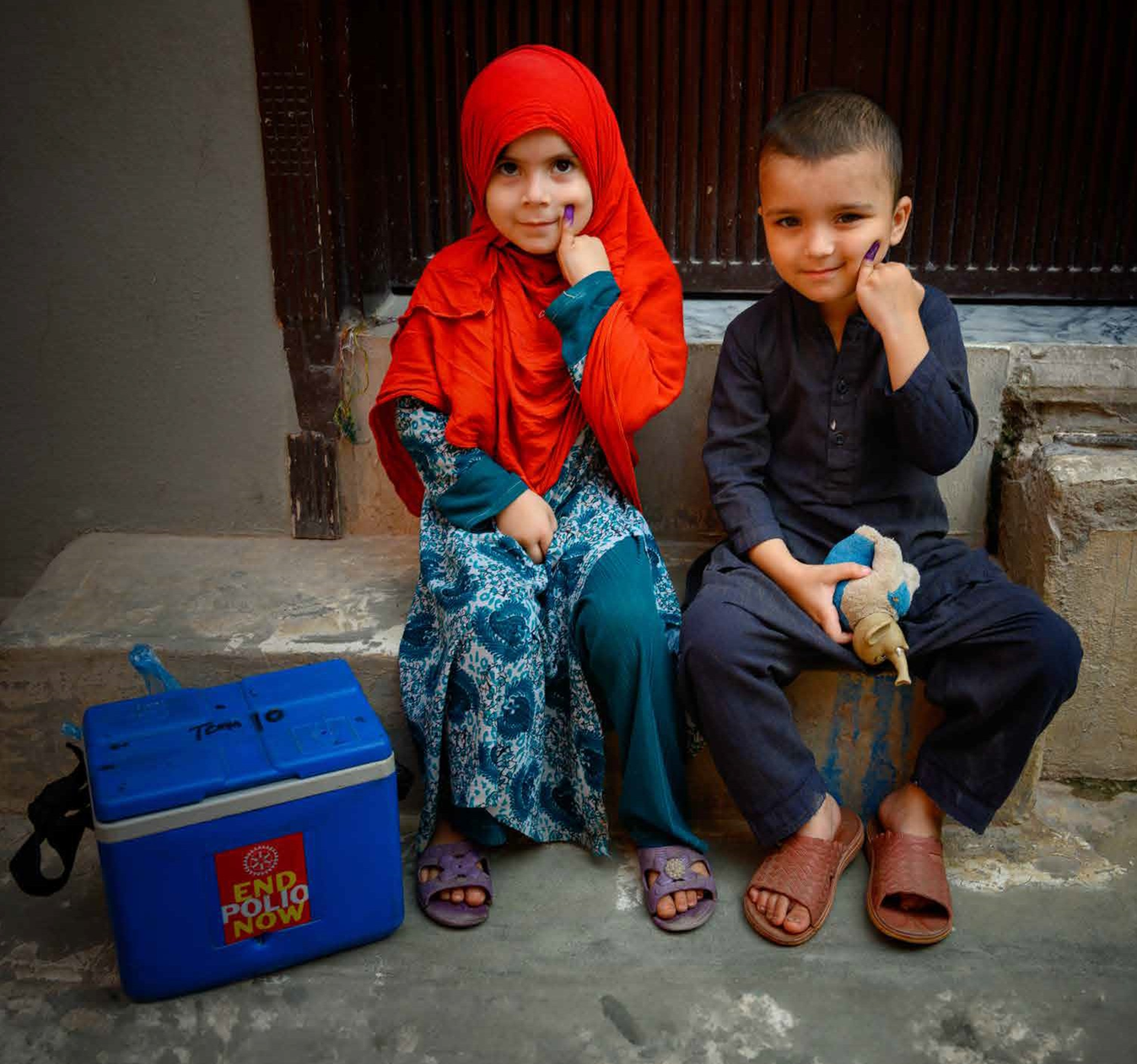 Two children smiling and showing vaccination marks, sitting next to a polio vaccine cold box.
