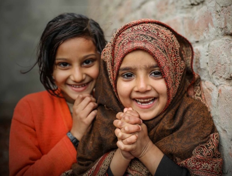 Two smiling girls, one in an orange sweater and the other in a patterned shawl, leaning against a brick wall.