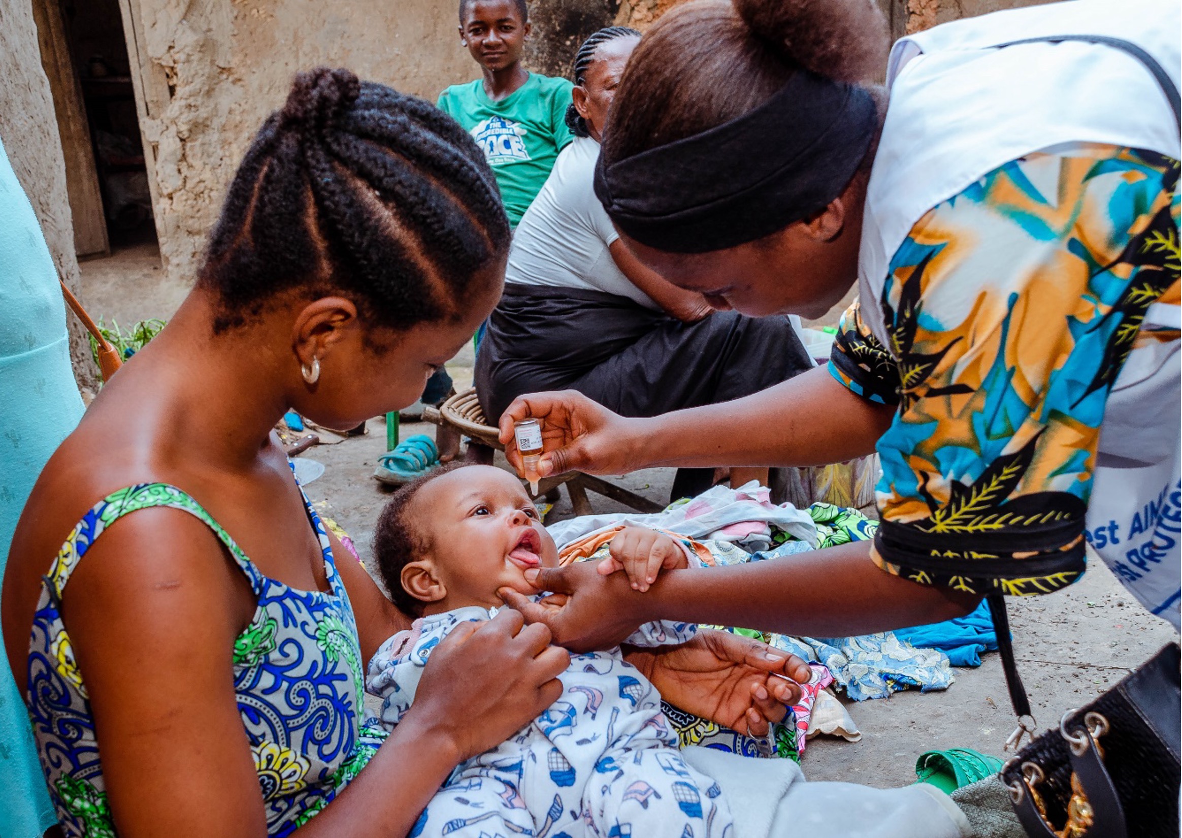 A health worker administers an oral polio vaccine to an infant held by their mother, while community members observe in a rural setting.