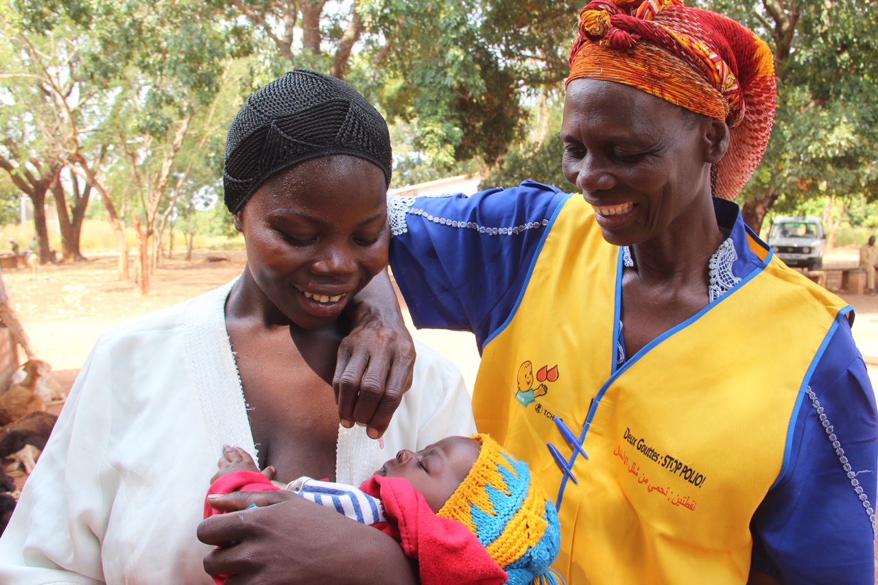 Two women smiling—one holding a baby, the other administering a vaccine