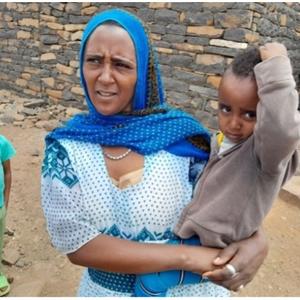 A mother holds her child close after receiving a vaccination, with another young sibling standing nearby. The family is pictured outside their home, illustrating the community's participation in vaccination efforts to protect children’s health.