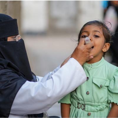 A health worker in a niqab administers an oral vaccine to a young girl wearing a green dress. The setting appears to be outdoors.