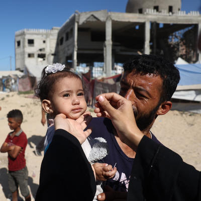A health worker in Gaza administers the nOPV2 vaccine to a child amid the devastation of ongoing conflict, embodying resilience and hope in the fight against polio.