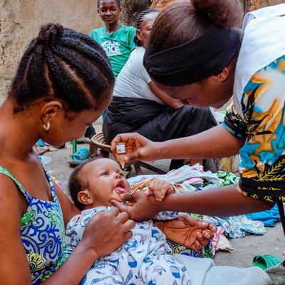 A health worker administers an oral polio vaccine to an infant held by their mother, while community members observe in a rural setting.