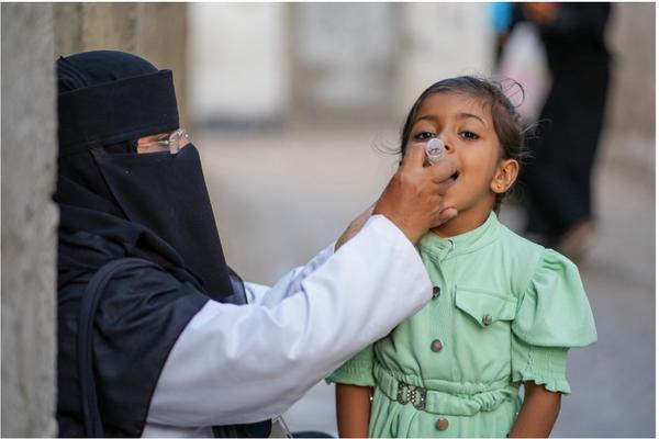 A health worker in a niqab administers an oral vaccine to a young girl wearing a green dress. The setting is outdoors.