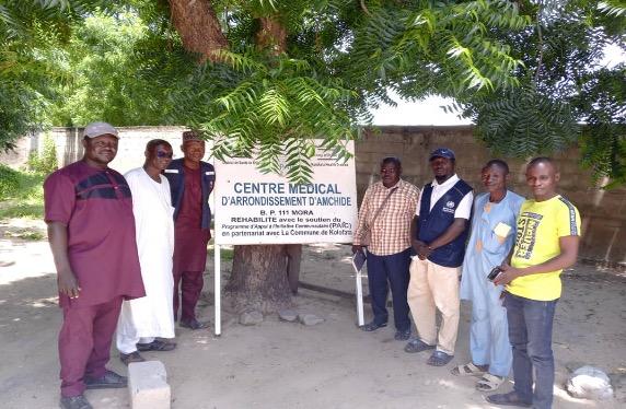 Group of health workers and community members standing together in front of a sign for the Centre Médical d’Arrondissement d’Anchidé, under a large tree.