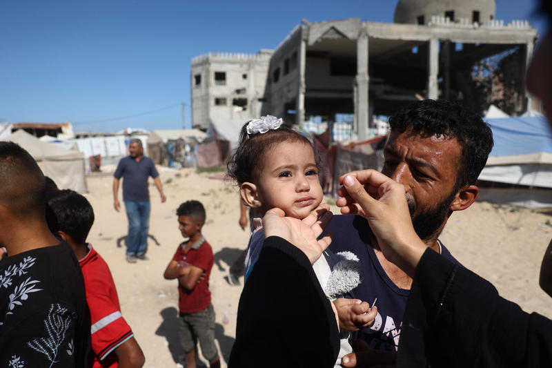 A health worker in Gaza administers the nOPV2 vaccine to a child amid the devastation of ongoing conflict, embodying resilience and hope in the fight against polio.