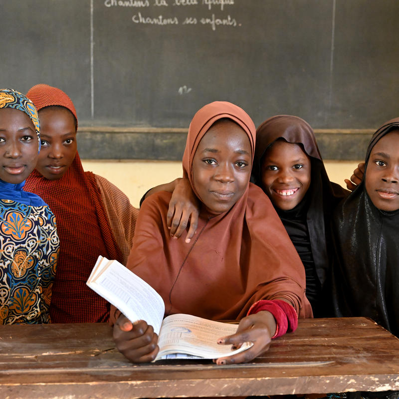 Un groupe de filles nigériennes en uniforme scolaire sont assises à un bureau et lisent un livre, souriant à la caméra. Un tableau noir est visible en arrière-plan.