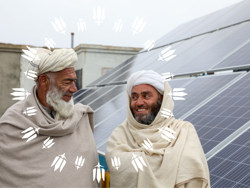 Two men wearing turbans and wool shawls stand together outdoors, smiling, with a large solar panel installation behind them.