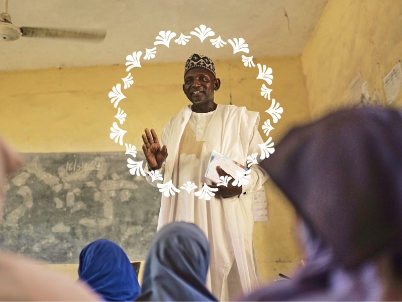 A man in a white robe and cap gestures while speaking at the front of a classroom, seen from behind students wearing headscarves.