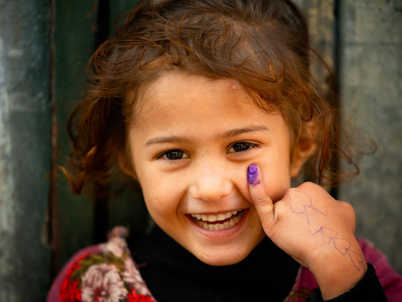 A young child looks directly at the camera smilling and showing her marked finger as an indicator that she took the polio vaccine.