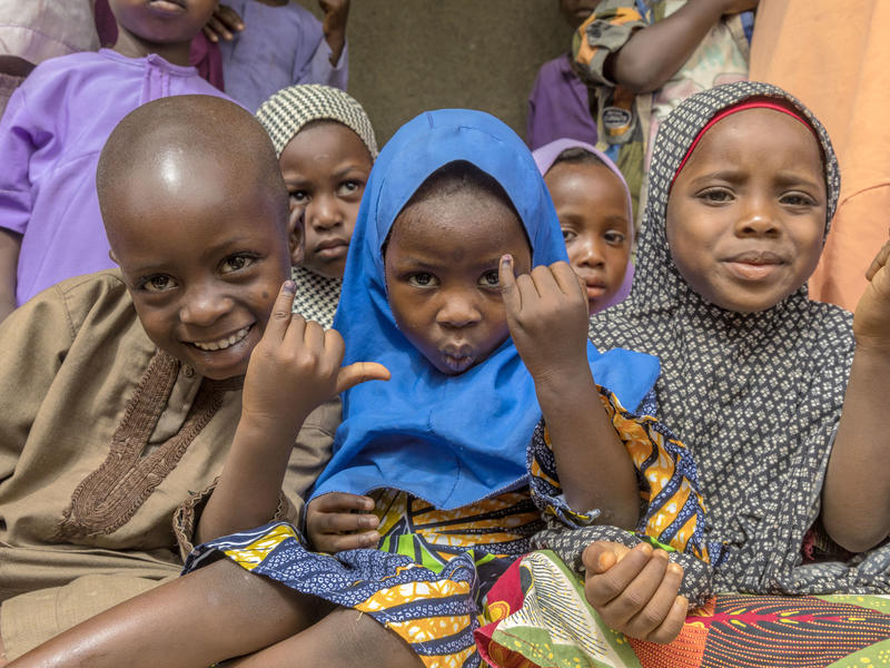 A group of Nigerian children sit close together indoors, several holding up fingers marked with purple ink and smiling at the camera.