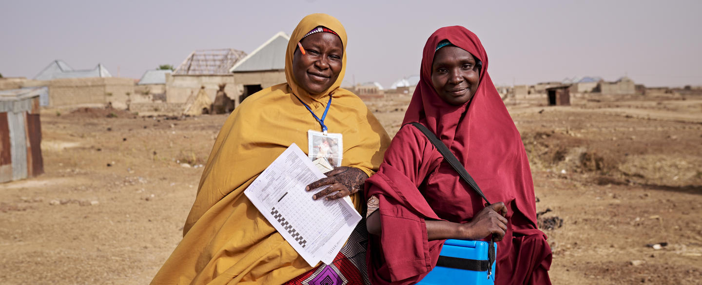 Two women wearing colorful hijabs stand in a dusty rural setting, holding a paper checklist and a small cooler used for medical supplies.