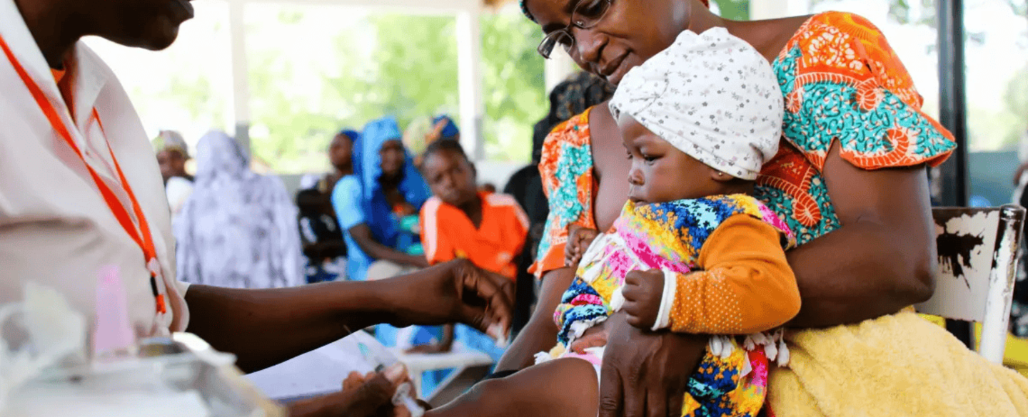 A health worker vaccinates a baby during an immunization session in Cameroon, while the mother, dressed in colourful traditional attire, holds the child on her lap. Other women wait in the background.