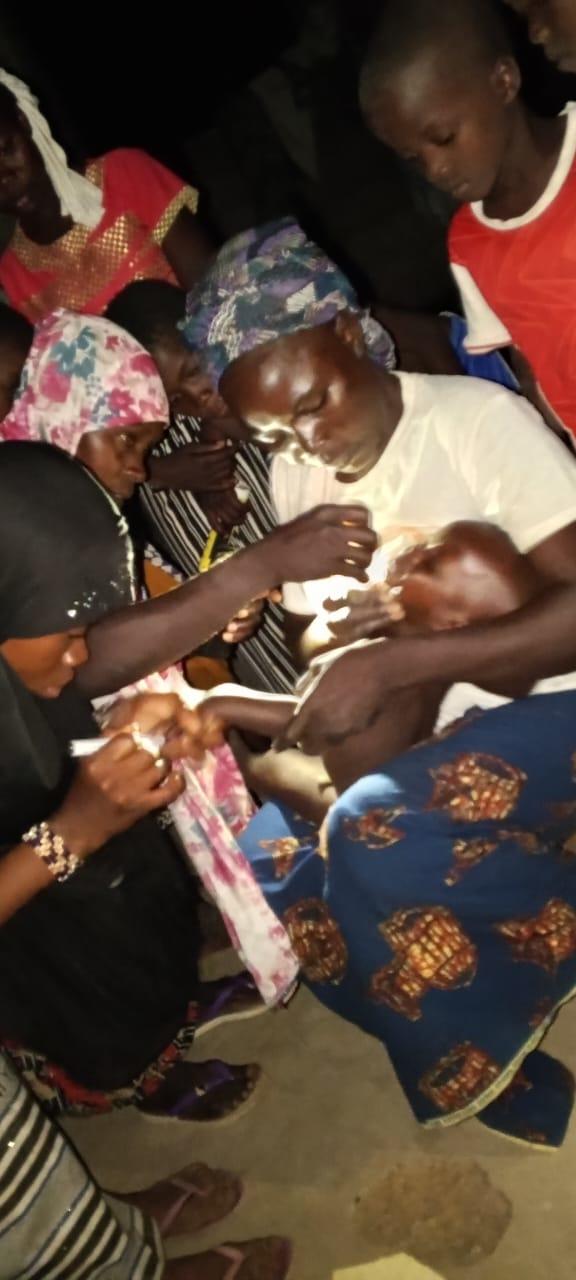 A nighttime vaccination scene in Zabré, Burkina Faso. A grandmother in a white t-shirt and blue headscarf holds a young infant while a health worker administers oral polio vaccine drops. A flashlight illuminates the scene as other family members, including women and children, gather around in the darkness.