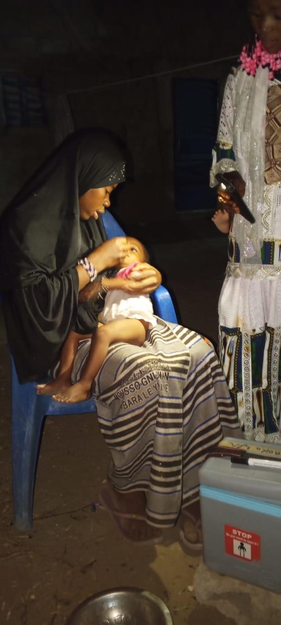 Nighttime polio vaccination in Zabré. A young mother in a dark hijab sits on a blue plastic chair, holding her infant who is receiving oral polio drops. A woman in a white lace dress stands nearby while a vaccine cold box is visible on the ground. The scene is lit by artificial light in the darkness.