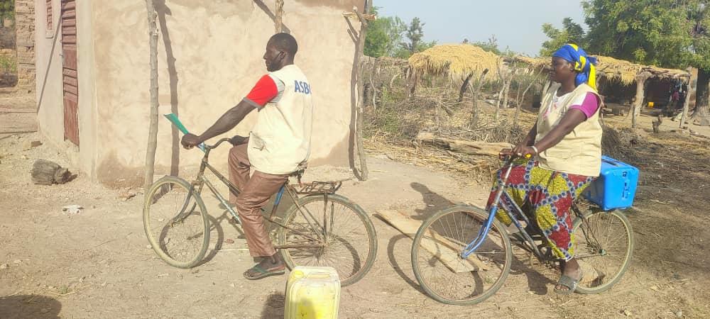 Two community health workers pause on their bicycles in a rural village in Nouna, Burkina Faso. A woman wearing a colorful patterned skirt and yellow headscarf carries a blue vaccine cold box on her bicycle. Her male colleague in an ASBC t-shirt holds documentation on his handlebars. Thatched-roof structures and agricultural storage are visible in the background.
