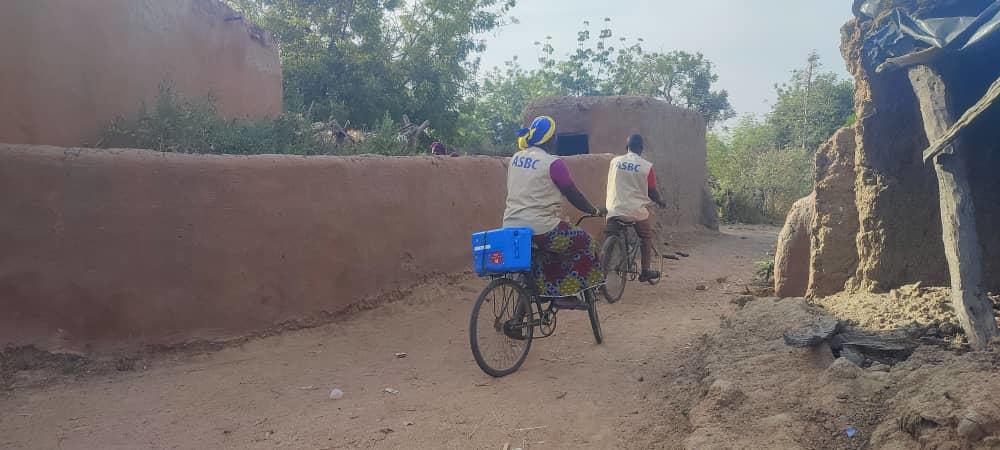 Two community health workers wearing ASBC vests ride bicycles side by side down a sandy path between traditional mud-walled compounds in Nouna, Burkina Faso. One carries a blue vaccine cold box on the back of their bicycle. The dusty road stretches ahead into a rural village landscape.