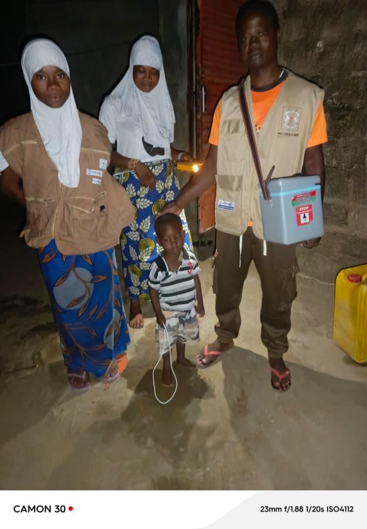 A vaccination team poses at night in Zabré. Two women in white hijabs and beige vests stand with a male health worker carrying a blue vaccine cold box. A young boy in a striped shirt stands in front of them. One woman holds a flashlight that illuminates the scene.